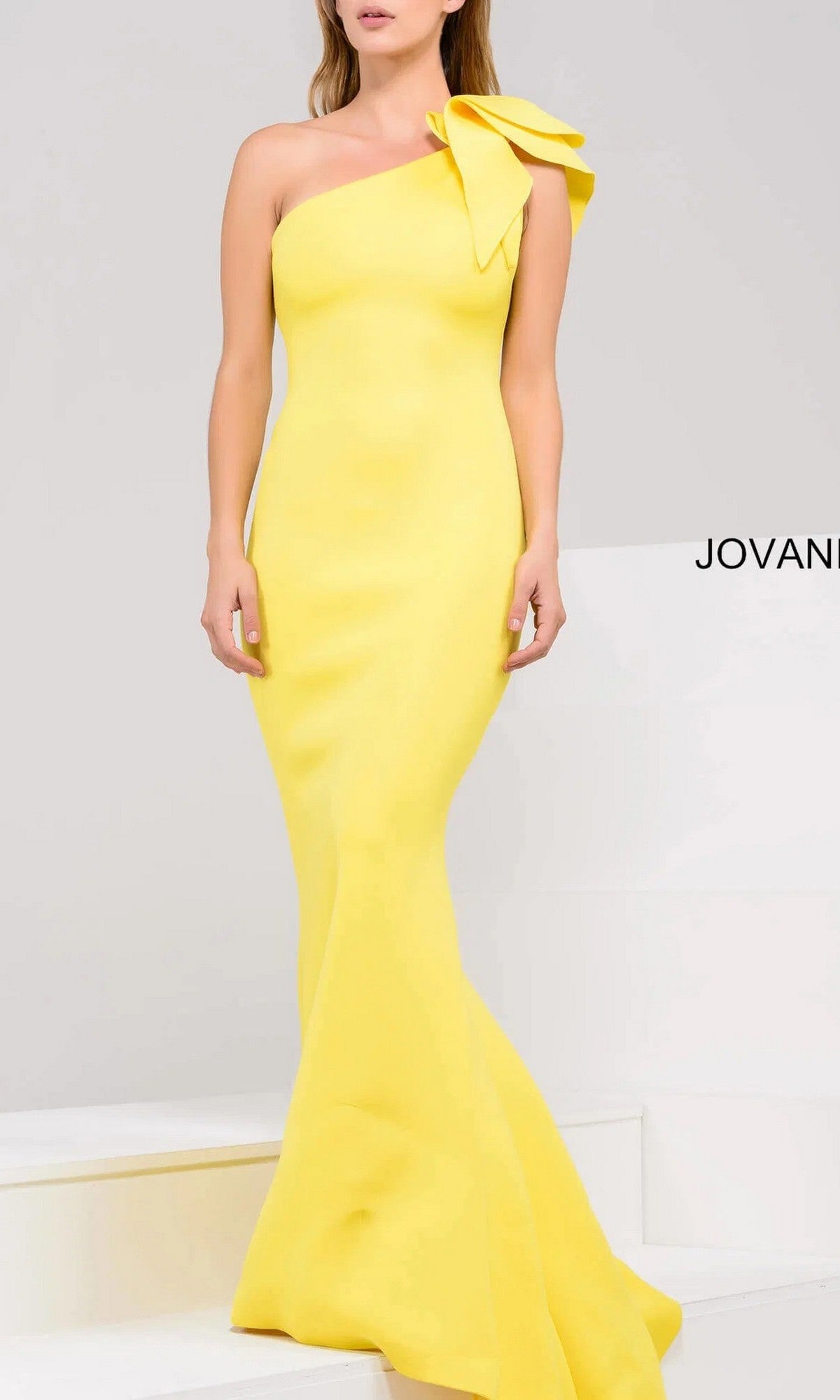 A woman stands elegantly on white steps in the Jovani 32602 One-Shoulder Mermaid Prom Gown, a fitted, floor-length yellow dress featuring a large bow on the shoulder.